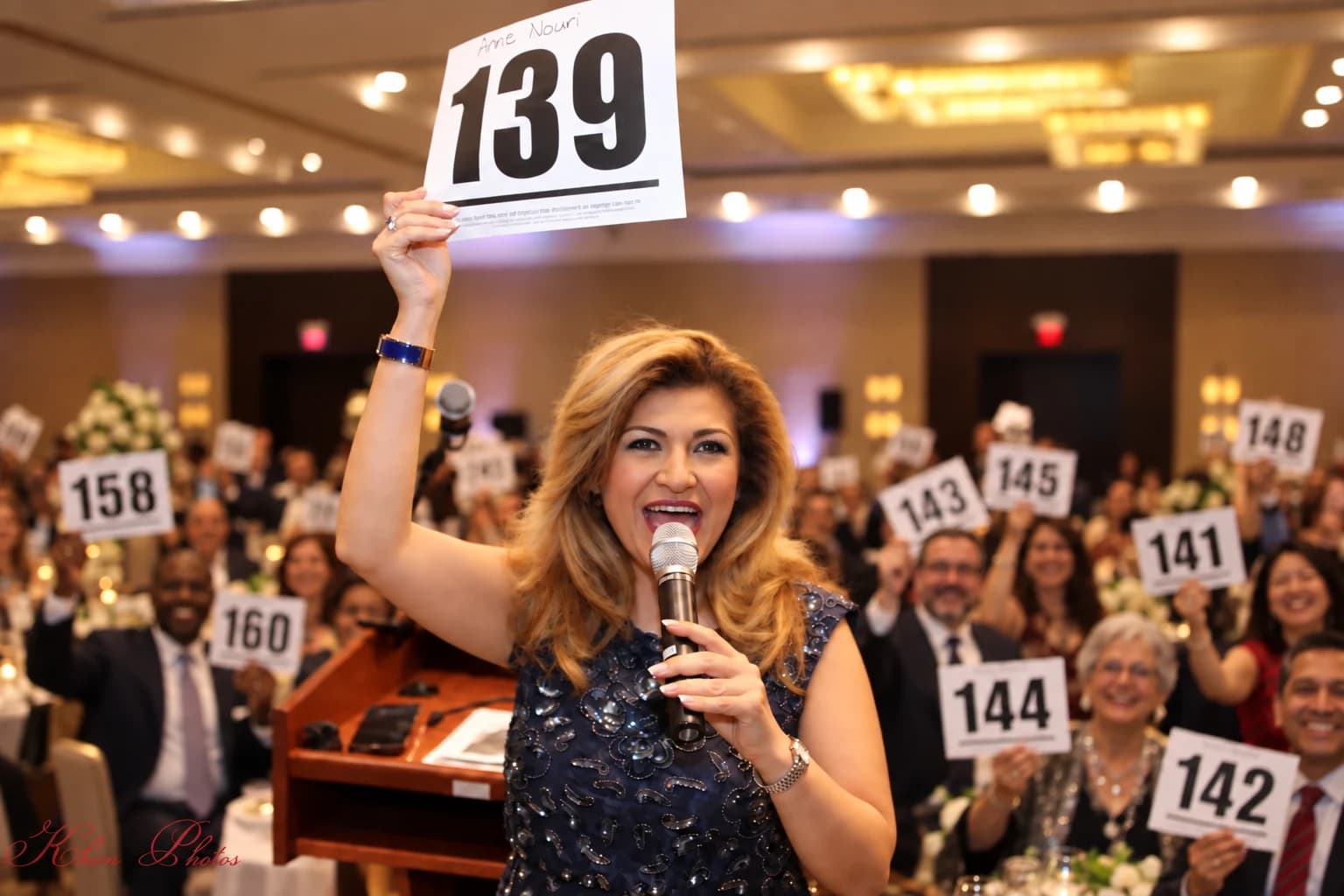 Illustrative benefit auction scene of Anne Nouri with bidders raising numbered paddles